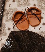 Pair of brown leather baby shoes on a patterned fabric background with 'Made in the USA' label.