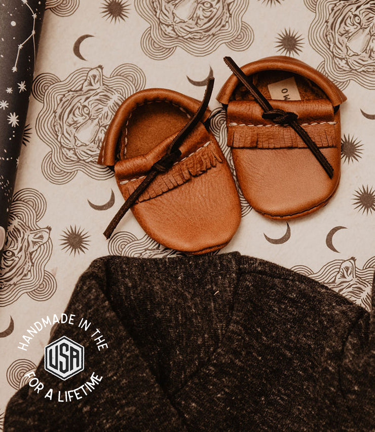 Pair of brown leather baby shoes on a patterned fabric background with &