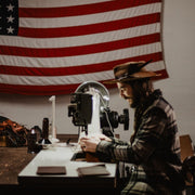 Blaine in the workshop making slim leather wallet at the sewing machine with American flag in the background