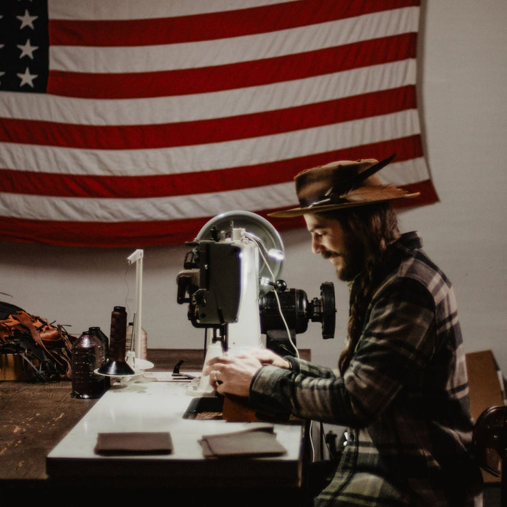 Blaine in the workshop making slim leather wallet at the sewing machine with American flag in the background
