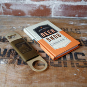 Book titled 'Beer Snob' and bottle opener on a wooden surface with a brick wall background