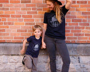 Home of the Lakers Water Tower Tee navy blue with light gray graphic  in front of a brick wall.