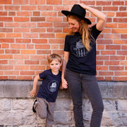 Woman and child wearing a navy t-shirts with a graphic design of a water tower that reads home of the lakers in front of a brick wall. 