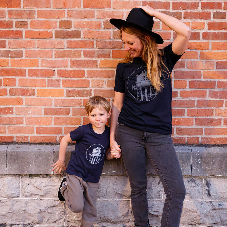 Woman and child wearing a navy t-shirts with a graphic design of a water tower that reads home of the lakers in front of a brick wall. 