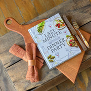 Cookbook titled 'Last Minute Dinner Party' on a wooden surface with cutlery and napkins.