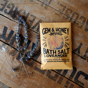 Yellow packet of Gem & Honey Infused Bath Salt Loveander on a wooden surface with lavender.
