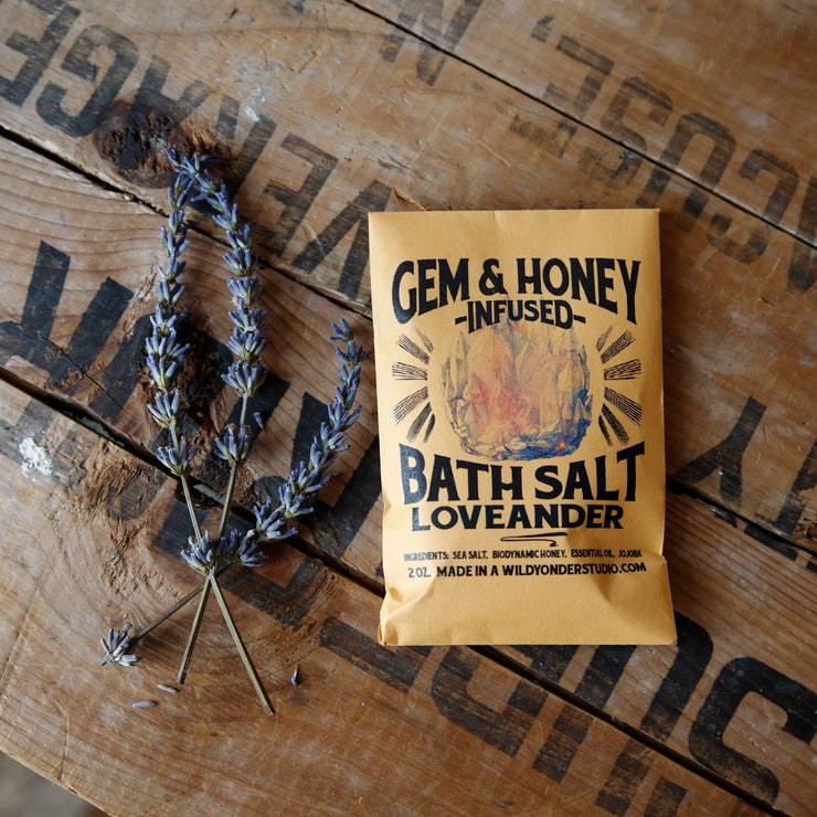 Yellow packet of Gem & Honey Infused Bath Salt Loveander on a wooden surface with lavender.