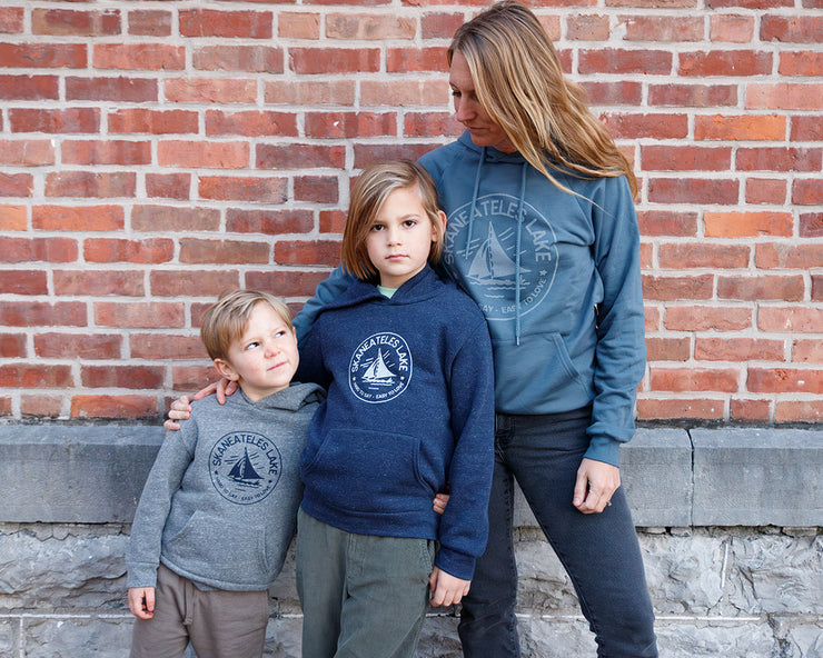 Woman and two children standing against a brick wall wearing matching Skaneateles Hard to Say Easy to Love sweatshirts by The Local Branch.