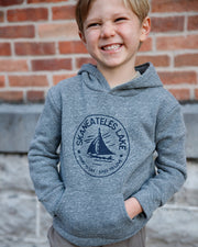 Child wearing a gray Skaneateles Hard To Say Easy to Love kids hoodie with sailboat design against a brick wall.