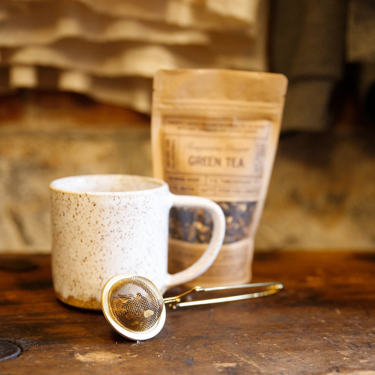 White ceramic mug with a wooden surface, green tea package, and tea infuser.