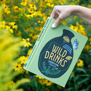 Hand holding a book titled 'Wild Drinks' with a natural background of greenery and yellow flowers