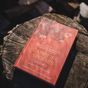 Book titled 'Campfire Stories Volume II' on a wooden stump with a campfire in the background