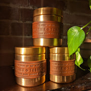 Three brass and leather candles stacked on a wooden surface with a brick wall background.